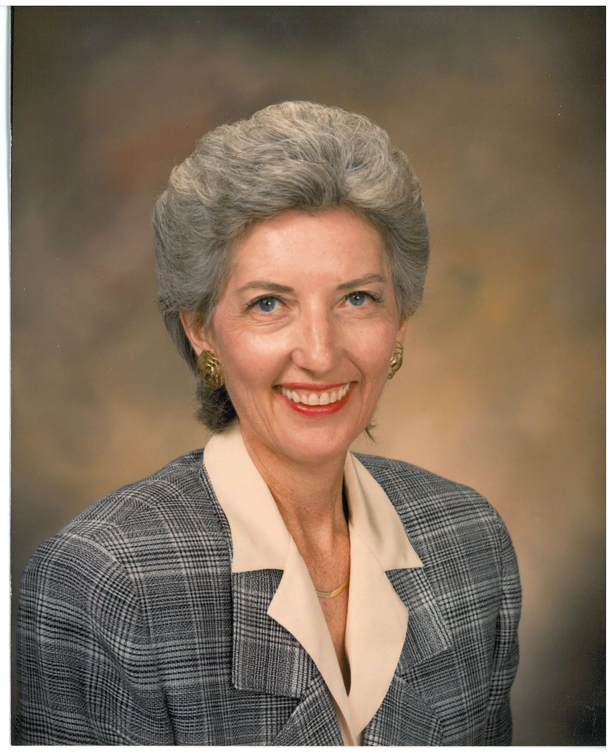 A woman with short gray hair, wearing a plaid blazer and a beige blouse, smiles against a neutral background.