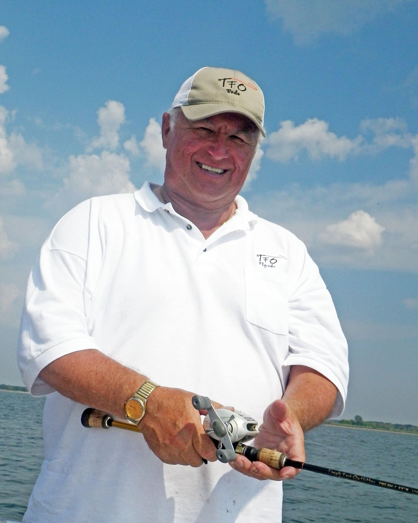 A man in a white shirt and beige cap is holding a fishing rod on a boat under a partly cloudy sky.